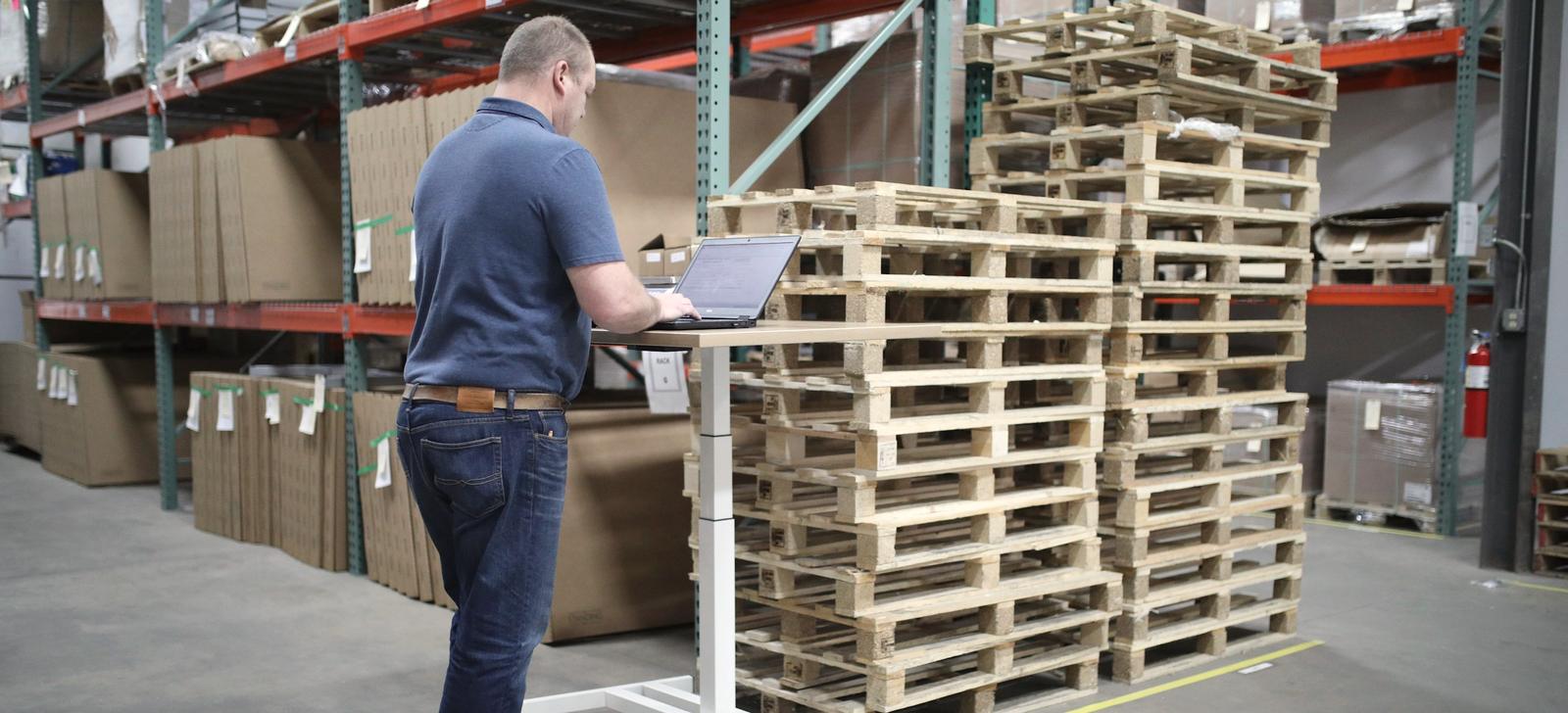 A man working at a laptop on a rolling stand in a warehouse aisle, beside stacked pallets.